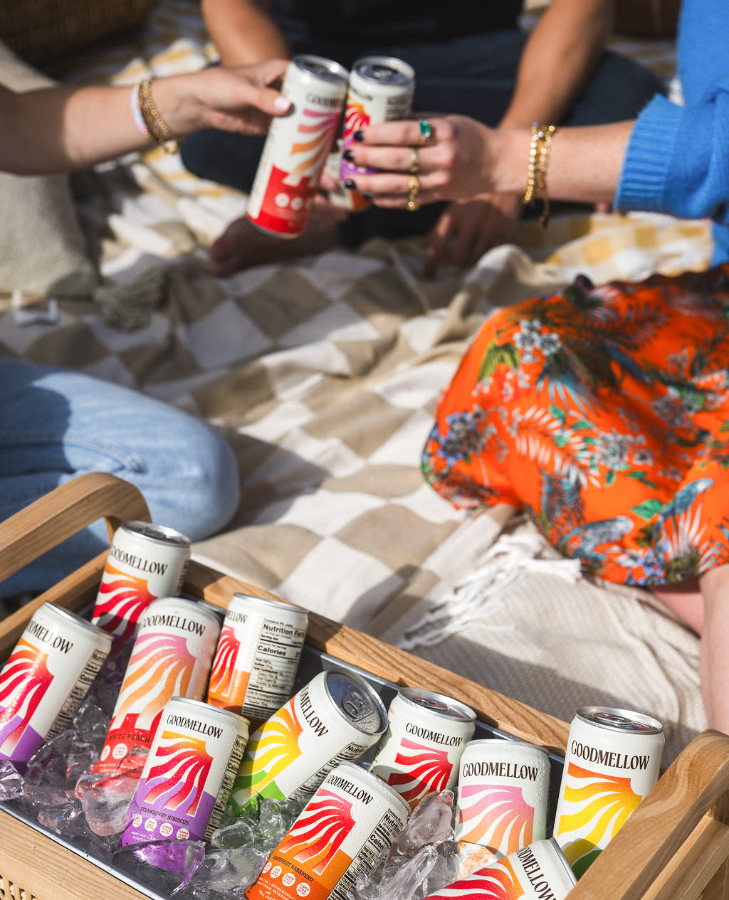 A wooden cooler filled with ice holds multiple cans of Livywell lemonade, with people sitting in the background clinking cans during an outdoor picnic.