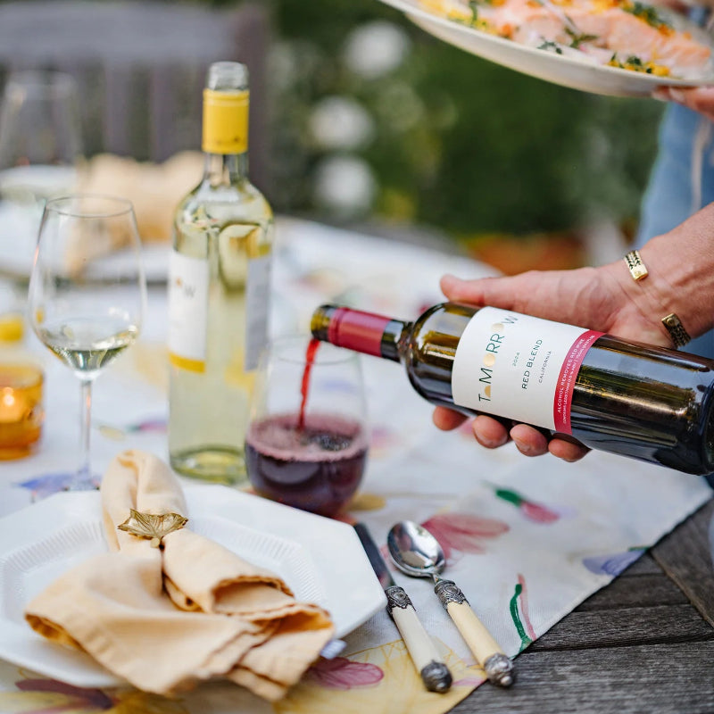Person pouring red wine into a glass at an outdoor dining table with food and wine bottles.